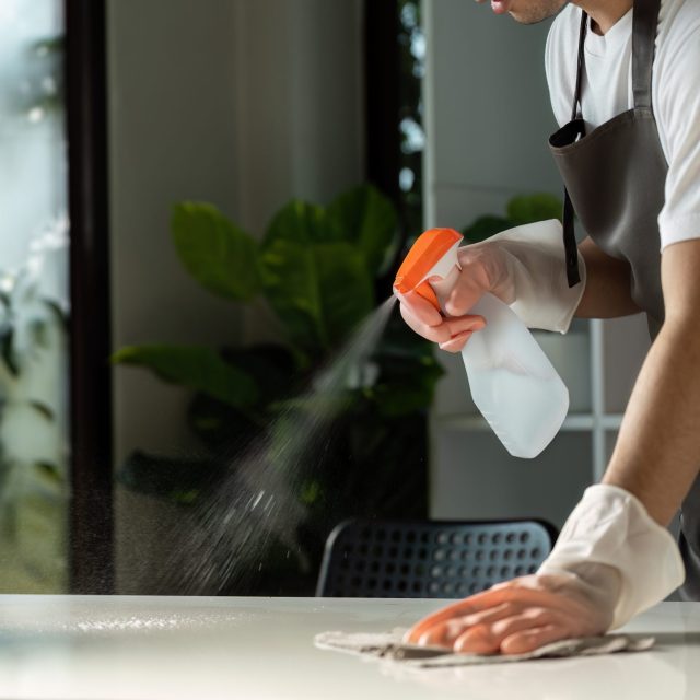 A person wearing gloves cleans a kitchen countertop with a spray bottle and cloth, emphasizing cleanliness and hygiene.