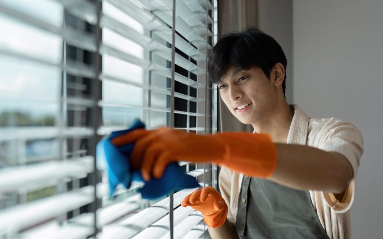 A housekeeper wearing gloves cleans window blinds, showcasing professional housekeeping skills in a modern home environment.