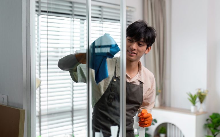 A young man wearing an apron and gloves cleans a glass door with a cloth and spray bottle in a bright, modern home interior.